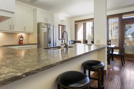 Modern kitchen with a marble countertop, stainless steel appliances, and bar stools. Dining area in background with large windows letting in natural light.