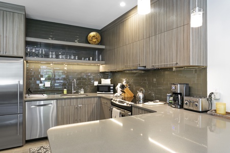 Modern kitchen with stainless steel appliances, grey cabinetry, and glossy dark green tile backsplash. Countertops feature various small kitchen appliances and utensils.