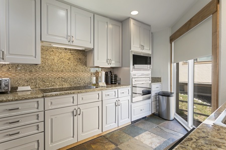 Modern kitchen with white cabinets, granite countertops, built-in appliances, a window with a shade, and a trash bin on a tiled floor.