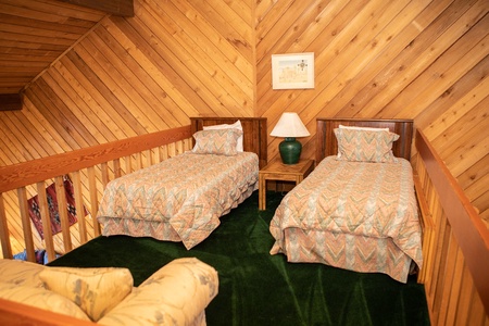 A loft bedroom with angled wooden walls, features two twin beds with patterned bedding, a green table lamp on a small wooden table, and green carpet flooring.