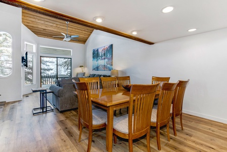 A dining area with a wooden table and eight chairs sits adjacent to a living room with a gray sofa and large windows overlooking a snowy scene. The ceiling has wooden beams and recessed lighting.