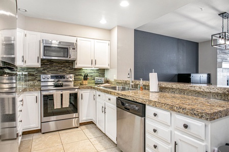 A modern kitchen with white cabinetry, stainless steel appliances, a granite countertop, a sink, and a dishwasher. The backsplash features a mosaic tile design, and the flooring is tiled.