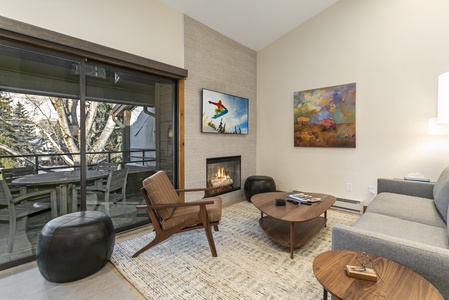 A modern living room features a gray sofa, wooden table, and armchair. A fireplace, a landscape painting, and a TV on the wall. A glass door opens to a balcony with outdoor seating and a view of trees.