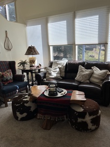 A living room with a leather sofa, cushions, a plaid armchair, a wooden coffee table with cowhide stools, and a side table with a lamp. Large windows with blinds let in natural light.