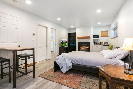 A studio apartment features a bed, wall-mounted TV, fireplace, small dining area, kitchenette, and a door leading to another room. The room is well-lit with a cozy, minimalist design.