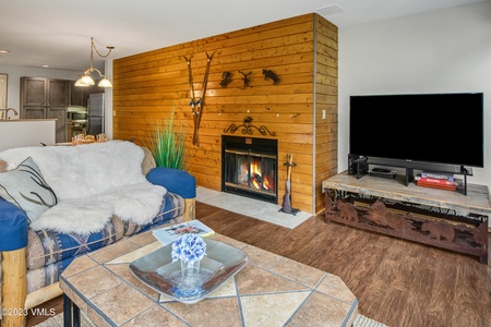 A cozy living room with a wooden accent wall, fireplace, and flat-screen TV. A couch with a fur throw is in the foreground, and a tile coffee table has a small centerpiece and magazine.