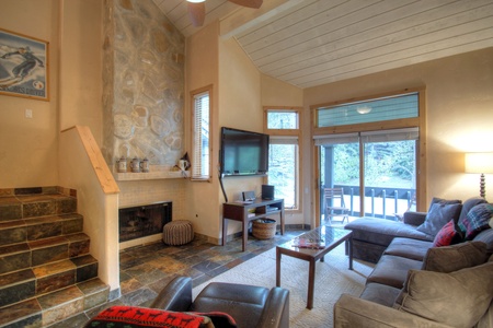 A cozy living room with a stone fireplace, L-shaped sofa, wall-mounted TV, and large windows leading to a balcony. Stairs are on the left, and a lamp illuminates the corner of the room.