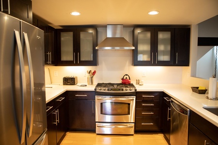 A modern kitchen features dark wood cabinets, stainless steel appliances including a refrigerator, oven, and range hood, and a countertop with utensils and a red kettle.