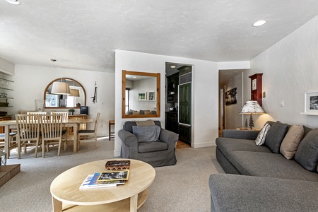 A living room with a round coffee table, gray sofa, and armchair. There's a mirror, dining table with chairs, and a wall cabinet. The area is well-lit with white walls and recessed lighting.