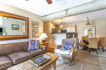 A living room with a brown leather sofa, a patterned armchair, a coffee table, and a lamp. The space extends into a kitchen and dining area with modern appliances, a dining table, and chairs.