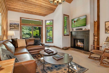 A cozy living room features a brown leather sectional sofa, a fireplace, a flatscreen TV mounted above the fireplace, a circular coffee table, wooden ceiling beams, and large windows with green views.