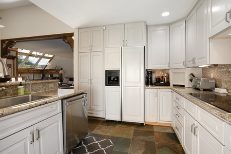 Modern kitchen with white cabinets, stainless steel appliances, granite countertops, and slate tile flooring. Natural light from large windows highlights the open layout.