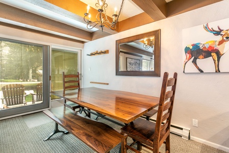 A wooden dining table with bench seating and high-back chairs is placed under a chandelier. A large mirror and colorful deer painting hang on the wall. Sliding glass doors lead to an outdoor patio.