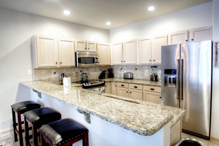 A modern kitchen with granite countertops, stainless steel appliances, light wood cabinets, and three black leather stools at the bar. The kitchen is well-lit with recessed lighting.