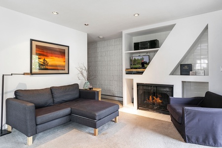 A modern living room features a dark gray sectional sofa and armchair, a fireplace with a TV above it, built-in shelves, a floor lamp, and framed artwork on the wall.