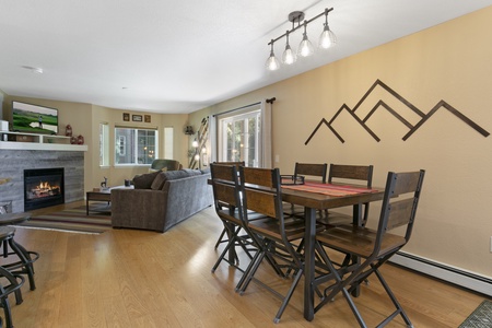 A dining area with a wooden table and chairs is adjacent to a living room featuring a fireplace, sofa, and wall-mounted TV. Wooden flooring and mountain-themed wall decor are visible.