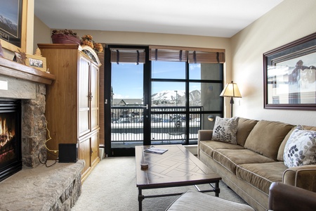 A cozy living room with a stone fireplace, wooden cabinets, a beige sofa with cushions, and a glass coffee table. Large windows offer a view of snow-covered mountains and a balcony.