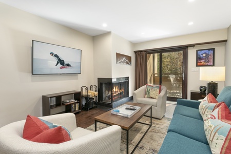 A cozy living room with a fireplace, TV showing a snowboarder, two sofas, a coffee table, and a sliding glass door leading to an outdoor balcony. Wall art and a lamp are also visible.