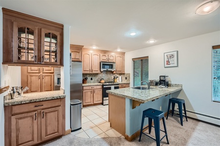 A modern kitchen with wooden cabinets, granite countertops, stainless steel appliances, and two blue stools at the breakfast bar. The kitchen has tile flooring and an open layout.