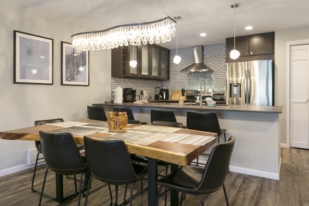 Modern kitchen and dining area with a wooden table, six black chairs, stainless steel appliances, and decorative lighting.