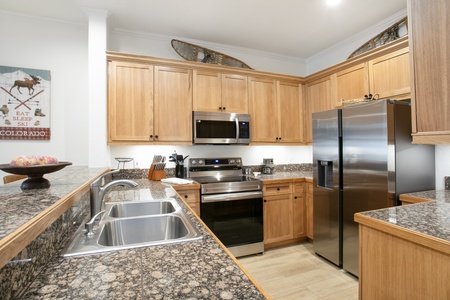 A modern kitchen features stainless steel appliances, including a stove and refrigerator, wooden cabinets, a dual sink, and a granite countertop, with decor items on top of cabinets.