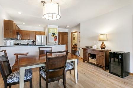 A dining area with a wooden table and four chairs, adjacent to a kitchen with wooden cabinets. A wall-mounted ski poster, a sideboard with a lamp, and a small refrigerator are also visible.