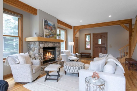 Cozy living room with stone fireplace, white sofas, two armchairs, and a round ottoman. Wooden beams and large windows create a warm atmosphere. Artwork above the mantel.