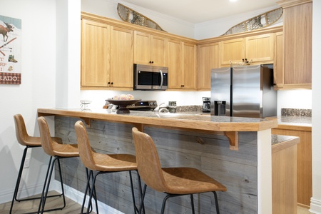 A modern kitchen features a wooden bar counter with four brown barstools, stainless steel appliances, light wood cabinets, and wall decor.