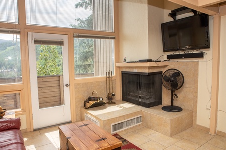 A cozy living room with a fireplace, a fan, wall-mounted TV, and a wooden coffee table. There is a door that leads to an outside balcony with a scenic view of trees and mountains in the background.