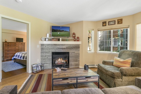 A cozy living room with a fireplace, television, sofa, and coffee table. The room has wooden floors, and a view of a bedroom through an open door. Several framed pictures are on the walls.