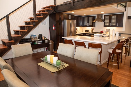 Modern kitchen with a central island featuring bar stools, stainless steel appliances, dark cabinets, and adjacent dining area. A staircase to the left leads to a loft.