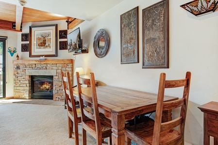 Cozy living room with a stone fireplace, wooden dining table, and chairs. The wall is decorated with framed art and a wall-mounted TV.