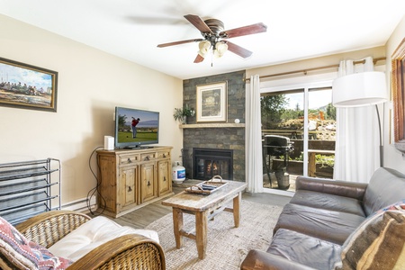 A cozy living room with a sofa, wicker chair, and wooden coffee table. A TV stands on a wooden cabinet near a fireplace, and a sliding door opens to an outdoor area with trees and greenery.