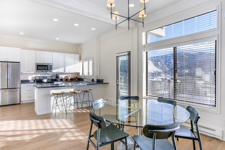 Modern kitchen and dining area with white cabinets, stainless steel appliances, a glass dining table with black chairs, large windows, and mountain views in the background.