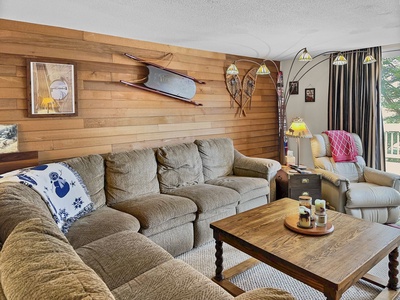 A cozy living room with a beige sectional sofa, a wooden coffee table, and a beige recliner. The wood-paneled wall features a mounted sled and decorative snowshoes.