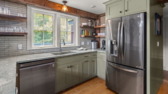 Modern kitchen with stainless steel appliances, green cabinets, and wooden shelves. The window above the sink provides natural light.