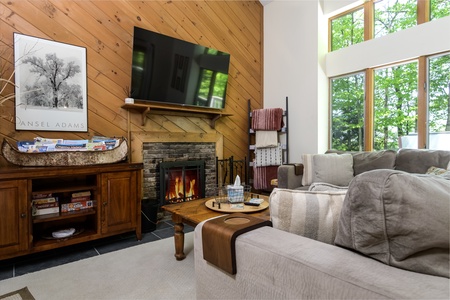 A cozy living room with a lit fireplace, large TV, gray sectional sofa, wooden coffee table, and blanket rack. A console holds a photo and games, while large windows provide views of greenery outside.