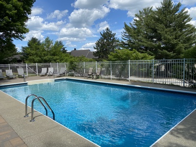 A fenced outdoor swimming pool with lounge chairs on a sunny day, surrounded by trees and houses in the background.