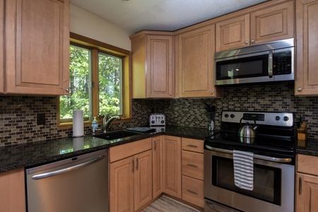 A modern kitchen features wooden cabinets, a stainless steel stove with a striped towel, a microwave, a dishwasher, a tiled backsplash, and a window overlooking greenery.