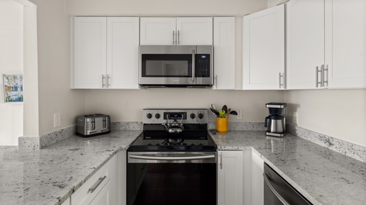 Modern kitchen with white cabinets, stainless steel appliances including a microwave, stove, toaster, and coffee maker. A yellow utensil holder and a kettle are on the counter.
