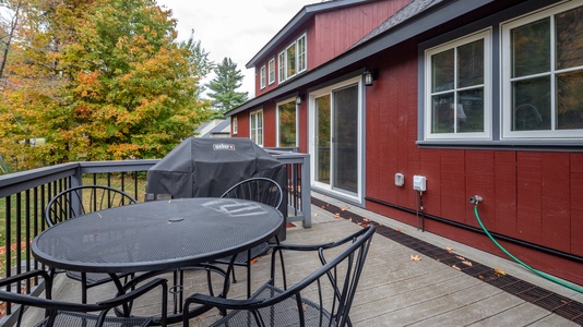 A wooden deck features a round metal table with chairs, a covered grill, and a red house with white-trimmed windows in the background. Trees with autumn foliage are visible on the left.