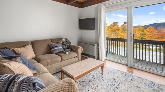 A cozy living room with a brown couch and a coffee table. A flat-screen TV is mounted on the wall. A sliding door opens to a balcony with a view of autumn foliage and distant hills.