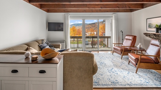 A cozy living room with a large L-shaped beige sofa, two brown leather armchairs, a TV mounted on the wall, a patterned blue and white rug, and a glass door opening to a balcony with a scenic view.