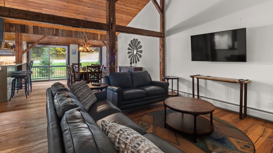 A cozy living room with a high wooden ceiling, leather sofas, a wall-mounted TV, a coffee table, and a dining area in the background with rustic decor and large windows.