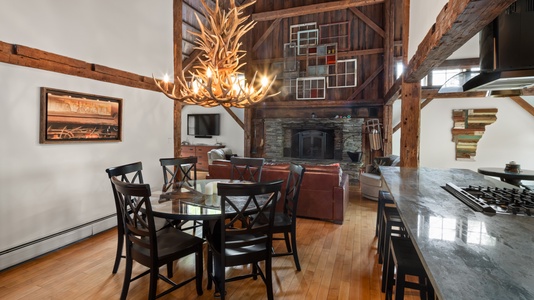 A rustic living and dining area features exposed wooden beams, a circular dining table with black chairs, a stone fireplace, a brown leather sofa, and a large antler chandelier.