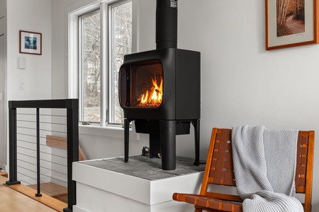 A black gas fireplace with a visible fire inside sits in a modern living space with white walls, a framed picture, a window, and a wooden chair with a gray blanket draped over it.