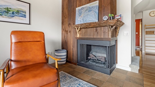 A cozy room features a brown leather chair, an electric fireplace with decor on the mantel, a basket, a wall map, and a framed picture. The floor has a tiled section by the fireplace and a blue rug.