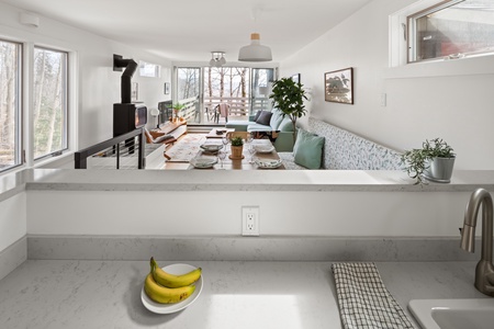 A modern, open-concept living and dining area with large windows, a set dining table, a grey sofa, a TV, and a wood stove. Two bananas are on a white kitchen counter in the foreground.