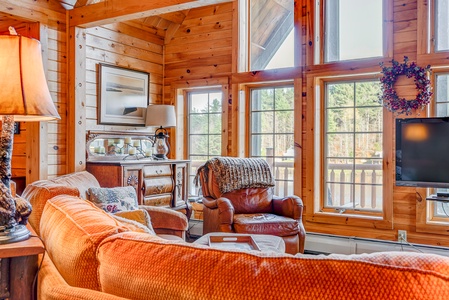 A cozy, sunlit living room with wooden walls, a large orange couch, an armchair, and a decorative wreath above the windows. A landscape painting and a lamp are placed on a wooden dresser.