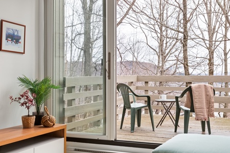 Living room with a sliding glass door opening to a wooden balcony. The balcony has two plastic chairs, a small table with a mug, and a brown blanket draped over one of the chairs. Trees are visible outside.